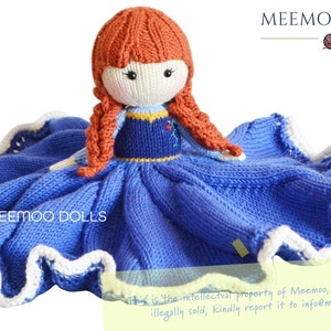 Pode incluir: Uma boneca de tricô com cabelo ruivo vestindo um vestido azul e um cobertor de tricô branco e azul. A boneca está sentada em um fundo branco. O texto "MEEMOO DOLLS" é visível no cobertor.