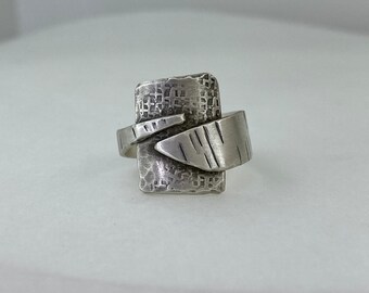 May include: A silver ring with a textured rectangular design and a smaller, textured, angled piece attached to the band.