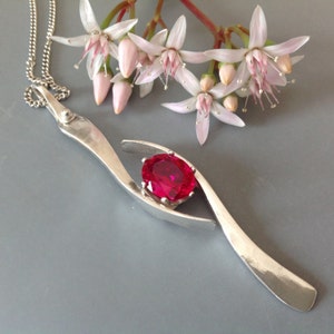 Puede incluir: Un collar de plata con un colgante que tiene una piedra preciosa roja brillante engastada en un engaste de plata. El colgante tiene forma de dos líneas curvas que se unen en la parte superior, creando un espacio para la piedra preciosa. El collar está en una cadena de plata.