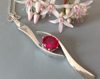 May include: A silver pendant necklace with a bright red gemstone set in a silver bezel. The pendant is shaped like two curved lines that meet at the top, creating a space for the gemstone. The necklace is on a silver chain.
