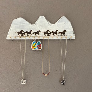 May include: White wooden jewelry holder shaped like a mountain range with a silhouette of horses running across the top. The holder has five hooks for hanging necklaces and earrings. There are three necklaces and a pair of earrings hanging from the hooks.