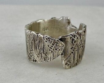 May include: A textured silver ring with a unique, open design. The ring features a bark-like pattern, creating a rustic and artistic aesthetic. The ring is a statement piece, perfect for adding a touch of individuality to any outfit.