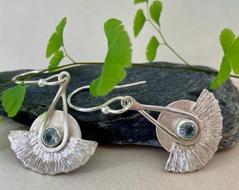 May include: A pair of silver earrings with a fan-shaped design and a round, light blue gemstone in the center. The earrings have a textured surface and a hook closure. Green leaves are in the background.