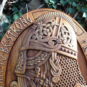 May include: A wooden carving of a Viking warrior's head wearing a helmet with chainmail. The carving is on a round wooden plaque with the words "TIME TO BE STRONG" and "RAGNAR" carved into the wood.