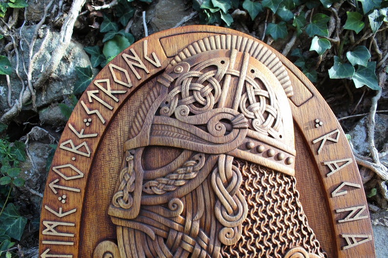 May include: A wooden carving of a Viking warrior's head wearing a helmet with chainmail. The carving is on a round wooden plaque with the words "TIME TO BE STRONG" and "RAGNAR" carved into the wood.