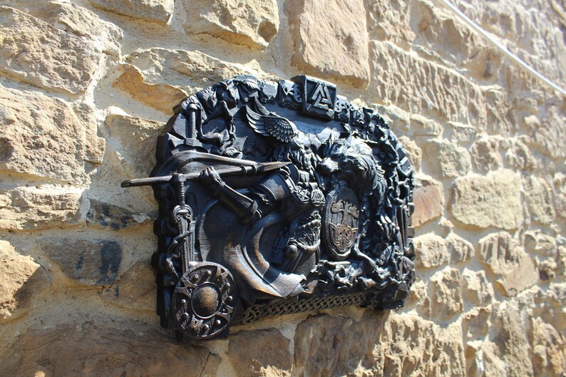 Puede incluir: Una placa de metal negro con una talla detallada de un guerrero que sostiene una lanza y un escudo. La placa est&aacute; montada en una pared de piedra y presenta un escudo de armas con las letras "A" y "S" entrelazadas.