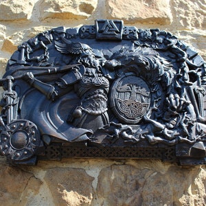 Puede incluir: Una talla de madera negra de un guerrero con un escudo y un lobo. La talla est&aacute; montada en una pared de piedra. El guerrero lleva armadura y un casco con alas. El lobo est&aacute; gru&ntilde;endo y muestra los dientes. El escudo tiene un dise&ntilde;o de nudo celta.