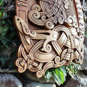 May include: A wooden wall hanging with intricate carvings of a knotwork design. The design features a central knot with swirling patterns and a stylized bird or animal figure. The wood is a rich brown color and the carving is detailed and precise.
