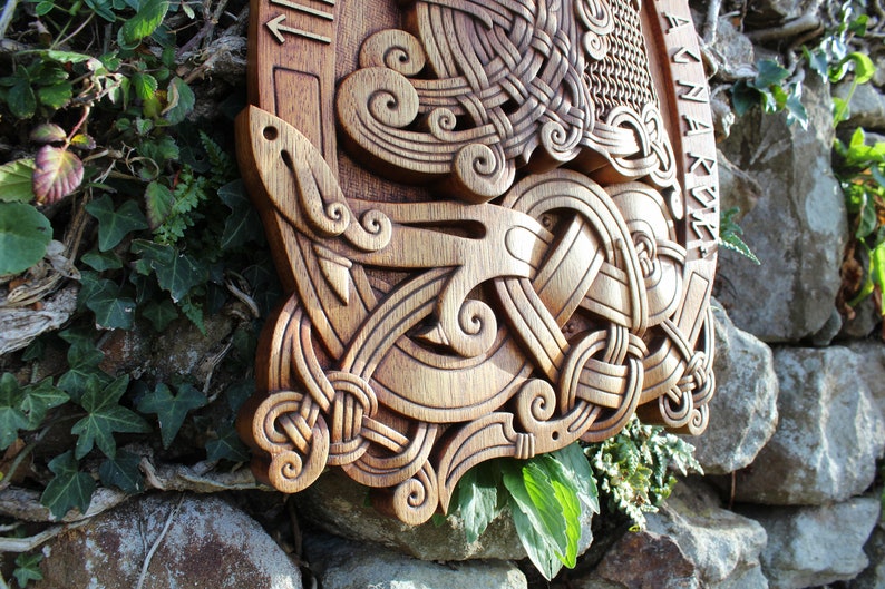 May include: A wooden wall hanging with intricate carvings of a knotwork design. The design features a central knot with swirling patterns and a stylized bird or animal figure. The wood is a rich brown color and the carving is detailed and precise.