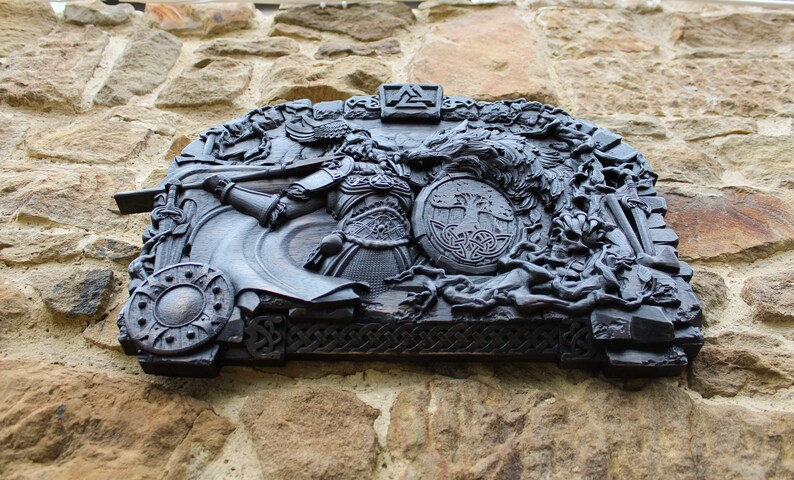 Puede incluir: Una talla de madera negra que representa a un guerrero, un lobo y un s&iacute;mbolo del &aacute;rbol de la vida. La talla est&aacute; montada en una pared de piedra.