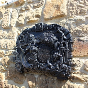 Puede incluir: Una placa de metal negro con una escultura en relieve detallada de un guerrero que sostiene una espada y un escudo, con un lobo y otras figuras en el fondo. La placa est&aacute; montada en una pared de piedra.