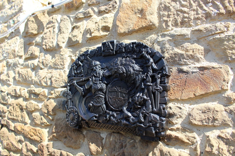 Puede incluir: Una placa de metal negro con una escultura en relieve detallada de un guerrero que sostiene una espada y un escudo, con un lobo y otras figuras en el fondo. La placa est&aacute; montada en una pared de piedra.