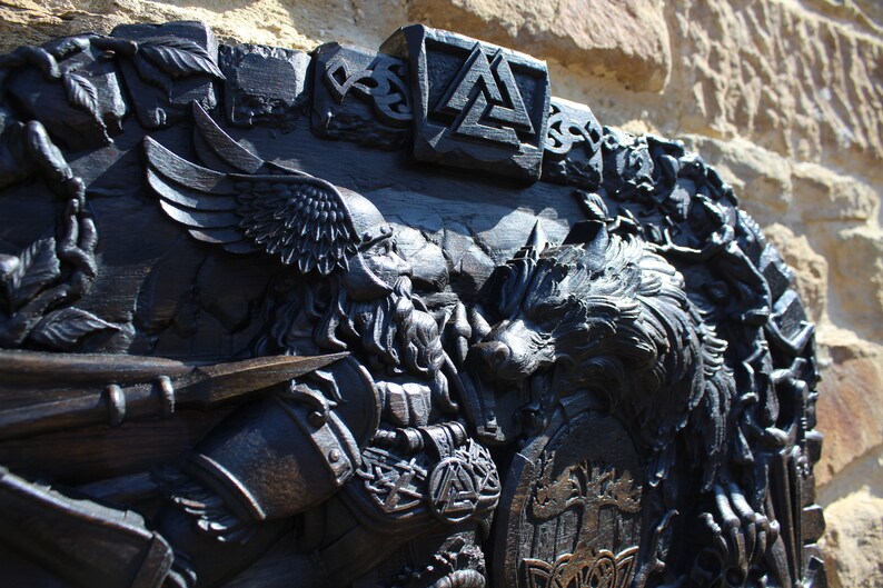 Puede incluir: Una talla de madera negra de un guerrero vikingo con un lobo, un escudo y un casco alado. La talla es detallada e intrincada, con una variedad de s&iacute;mbolos y dise&ntilde;os.