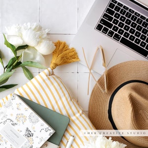 May include: A flat lay featuring a white and yellow striped pillow, a notebook with a tiger print, a green journal, a white pen, a straw hat, sunglasses, and a white peony flower.
