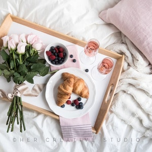 May include: A wooden tray with a white background, filled with a bouquet of pink roses, two croissants, a bowl of berries, and two glasses of champagne. The tray is on a white bed with a pink throw pillow.