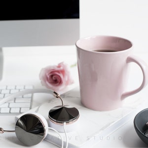 May include: White headphones with rose gold accents and black earcups, a pink mug with coffee, a white keyboard, and a pink rose on a white surface.