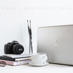 Pode incluir: Um computador portátil branco com um logotipo da Apple prateado na tampa, uma câmera preta com uma lente, uma xícara de café branca e uma pilha de cadernos em uma superfície branca. Há também dois pincéis em um vaso de vidro.