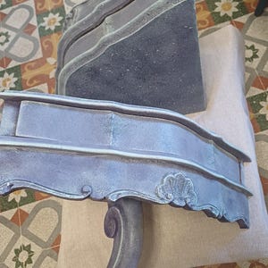 May include: Two-piece, ornate, gray-blue furniture set. The top piece is a rectangular block with layered edges. The bottom piece has a curved front with a decorative shell design and a scrolled leg. The pieces are on a patterned tile floor.