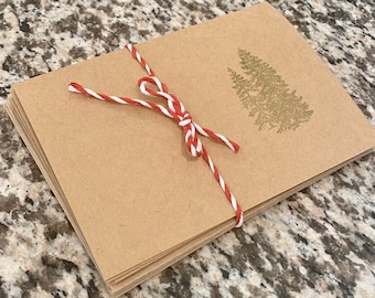 May include: A stack of kraft paper cards tied with red and white twine. A gold-colored illustration of two pine trees is printed on the top card. The cards are stacked neatly and tied with a bow.