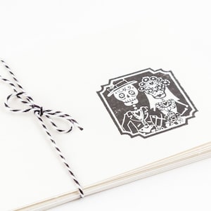 May include: A stack of cream-colored paper tied with black and white twine. A black and white illustration of a skeleton couple in formal attire is printed on the top sheet, framed by a decorative border. The image evokes a Day of the Dead theme.