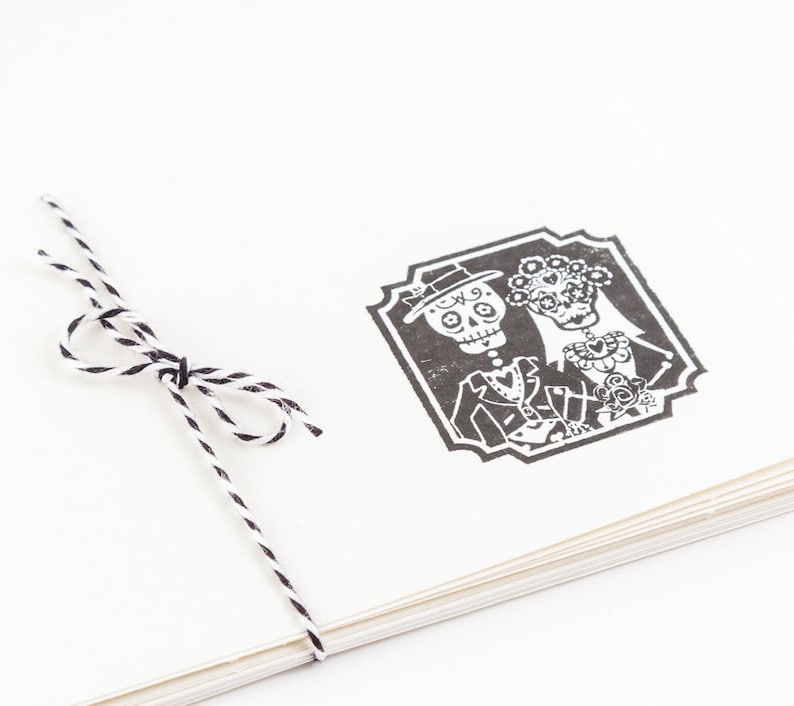 May include: A stack of cream-colored paper tied with black and white twine. A black and white illustration of a skeleton couple in formal attire is printed on the top sheet, framed by a decorative border. The image evokes a Day of the Dead theme.