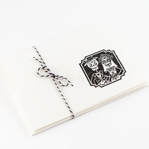 May include: A stack of white cards tied with black and white twine. The top card features a black and white illustration of a skeleton couple in formal attire, within a decorative frame. The design is reminiscent of Dia de los Muertos.