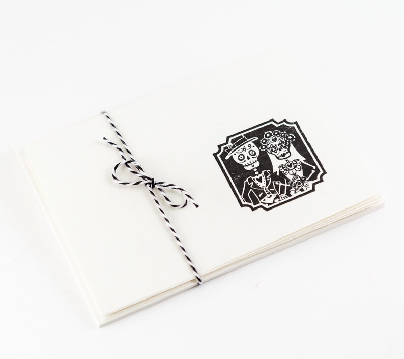 May include: A stack of white cards tied with black and white twine. The top card features a black and white illustration of a skeleton couple in formal attire, within a decorative frame. The design is reminiscent of Dia de los Muertos.