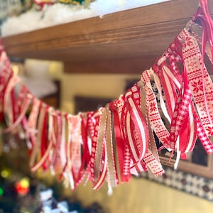 Może przedstawiać: Świąteczna girlanda złożona z czerwonych, białych i beżowych wstążek. Wstążki mają różne wzory, w tym płatki śniegu i paski. Girlanda zawieszona jest na rozmytym tle, sugerującym motyw świąteczny lub sezonowy.