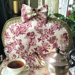 Könnte beinhalten: Ein Vintage-Teewärmer mit cremefarbenem und rotem Blumenmuster, das Engel und Rosen zeigt. Der Teewärmer hat eine dekorative Schleife und goldfarbene Verzierungen. Eine silberne Teekanne und eine Teetasse sind ebenfalls zu sehen.