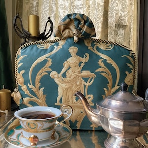 Könnte beinhalten: Eine teal- und goldfarbene dekorative Teewärmer mit klassischem Design, eine silberne Teekanne und ein Tassen- und Untertassenset auf einem Silbertablett. Die Tasse ist mit Tee gefüllt. Goldene Kerzen und ein Kerzenhalter sind im Hintergrund.