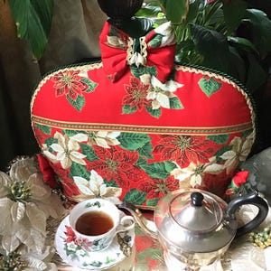 Könnte beinhalten: Rote und grüne Weihnachts-Teekanne mit einem Weihnachtsstern-Muster und einer goldenen Schleife. Die Teekanne ist herzförmig und hat einen grünen Rand. Im Hintergrund stehen eine silberne Teekanne und eine Teetasse auf einem silbernen Tablett.