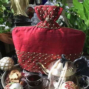 Peut inclure: Une housse à thé rouge avec des bordures de perles dorées et blanches. La housse est en forme de sac et a un pompon de perles sur le dessus. La housse est posée sur un plateau argenté avec une théière, des tasses à thé et d'autres objets décoratifs.