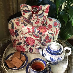 Könnte beinhalten: Eine florale Teekanne mit einer roten Samtschleife und silbernen Verzierungen. Die Teekanne steht auf einem silbernen Tablett mit einer Teekanne, einer Teetasse und Keksen.