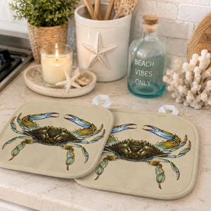 May include: Two beige square pot holders featuring a detailed illustration of a blue crab. The crab has blue claws and legs, with a brown body. The pot holders have white hanging loops.