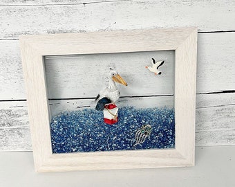 May include: A white wooden frame with a glass front containing blue sand-like material. A white bird with black accents is perched on a red and white buoy. A white seagull is flying above the bird. A blue jellyfish is in the sand.
