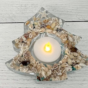Puede incluir: Un portavelas transparente en forma de estrella lleno de conchas marinas y una pequeña vela LED brillante. El soporte está decorado con varias conchas marinas pequeñas y una estrella de mar. El portavelas es un artículo decorativo.
