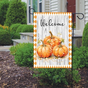 May include: A welcome garden flag with a white woodgrain background and orange diamond pattern border. The flag features three orange pumpkins with gold accents and the word "Welcome" in black lettering.