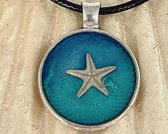 May include: A round pendant necklace with a starfish design. The pendant has a silver-coloured frame and a blue and teal background. The starfish is light beige. The necklace has a black cord.