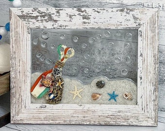 May include: A white wooden frame with a glass front containing a seashell and sand scene. The seashell is decorated with red, white, and blue accents. The frame is sitting on a white wooden surface.
