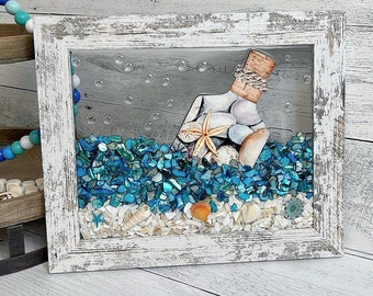 May include: A whitewashed wooden frame with a glass front containing a bottle with a cork stopper, seashells, and a starfish. The bottle is partially submerged in blue and white glass beads.