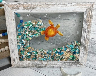May include: A framed artwork featuring a sea turtle swimming through a wave made of blue and green glass beads. The frame is whitewashed wood with a distressed finish. The artwork is signed "Oceanside".