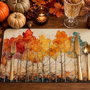May include: A rectangular placemat featuring a watercolor-style illustration of a forest with autumn foliage in shades of red, orange, yellow, and teal. A gold-toned fork, knife, and spoon are placed on the surface of the placemat.