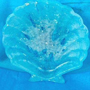 Può includere: Un piatto artigianale a forma di conchiglia, nei toni del blu e del bianco, con finitura lucida. Il piatto presenta una superficie strutturata con scaglie d'oro incorporate e accenti blu scuro, creando un'estetica unica ispirata all'oceano.
