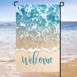 Puede incluir: Una bandera de jardín de bienvenida con una escena de playa. La bandera presenta un océano azul con olas blancas espumosas rompiendo en una playa de arena. La palabra "bienvenido" está escrita en cursiva azul sobre la arena.