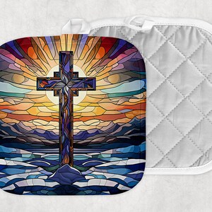 May include: A square pot holder with a colorful stained-glass design featuring a cross, sun, and landscape. The cross is purple and blue, set against a sunset of orange, yellow, and blue hues. A white quilted pot holder is next to it.