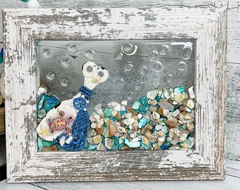 May include: A white framed picture featuring a blue and white sea turtle with a seashell on its back. The turtle is surrounded by a variety of seashells and glass beads in shades of blue, white, and brown.