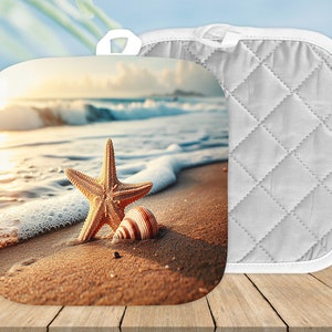May include: A pair of white quilted pot holders with a beach scene printed on one side. The scene features a starfish and a seashell on a sandy beach with ocean waves in the background.