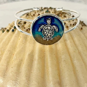 May include: Silver-tone bangle bracelet with a round charm featuring a detailed silver turtle against a blue and sand-colored background. The bracelet is displayed on a textured, light-colored surface, evoking a beach theme.