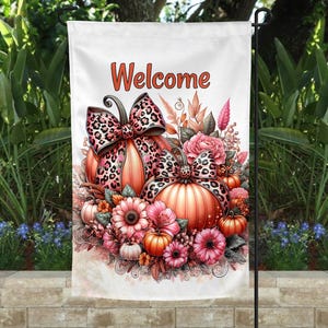 以下が含まれることがあります： オレンジ色のスクリプトで「Welcome」という言葉が書かれた白いガーデンフラッグ。オレンジ、ピンク、茶色の色合いのカボチャ、花、葉のデザインが特徴です。カボチャにはヒョウ柄のリボンが付いています。