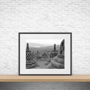 Op de afbeelding: Zwart-wit ingelijste foto van het Borobudur-tempelcomplex in Indonesië. De afbeelding toont verschillende stupa's en het omliggende landschap. De foto wordt weergegeven op een houten plank tegen een witte bakstenen muur.